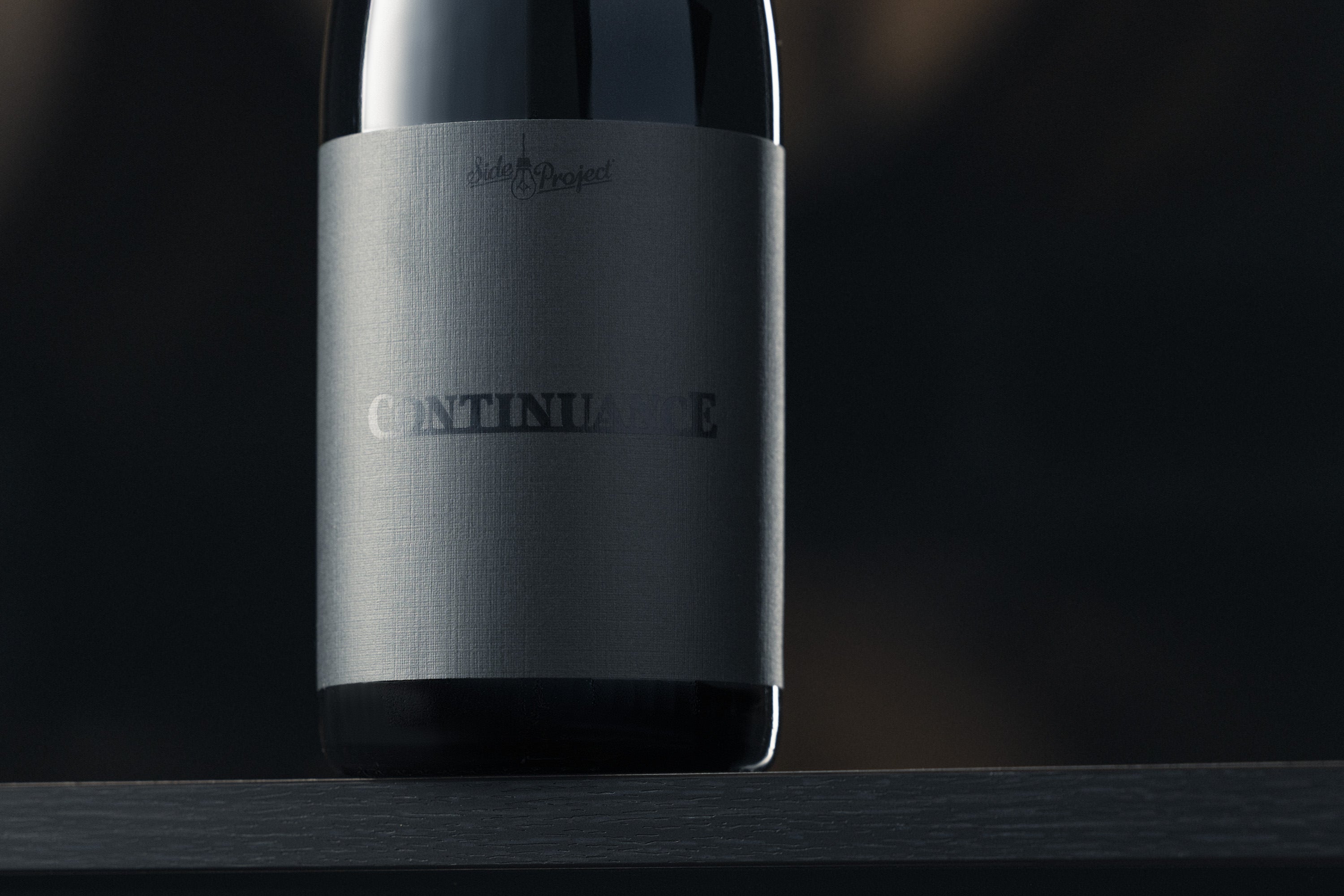 Beer bottle with 'Continuance' label on a dark background in front of barrels