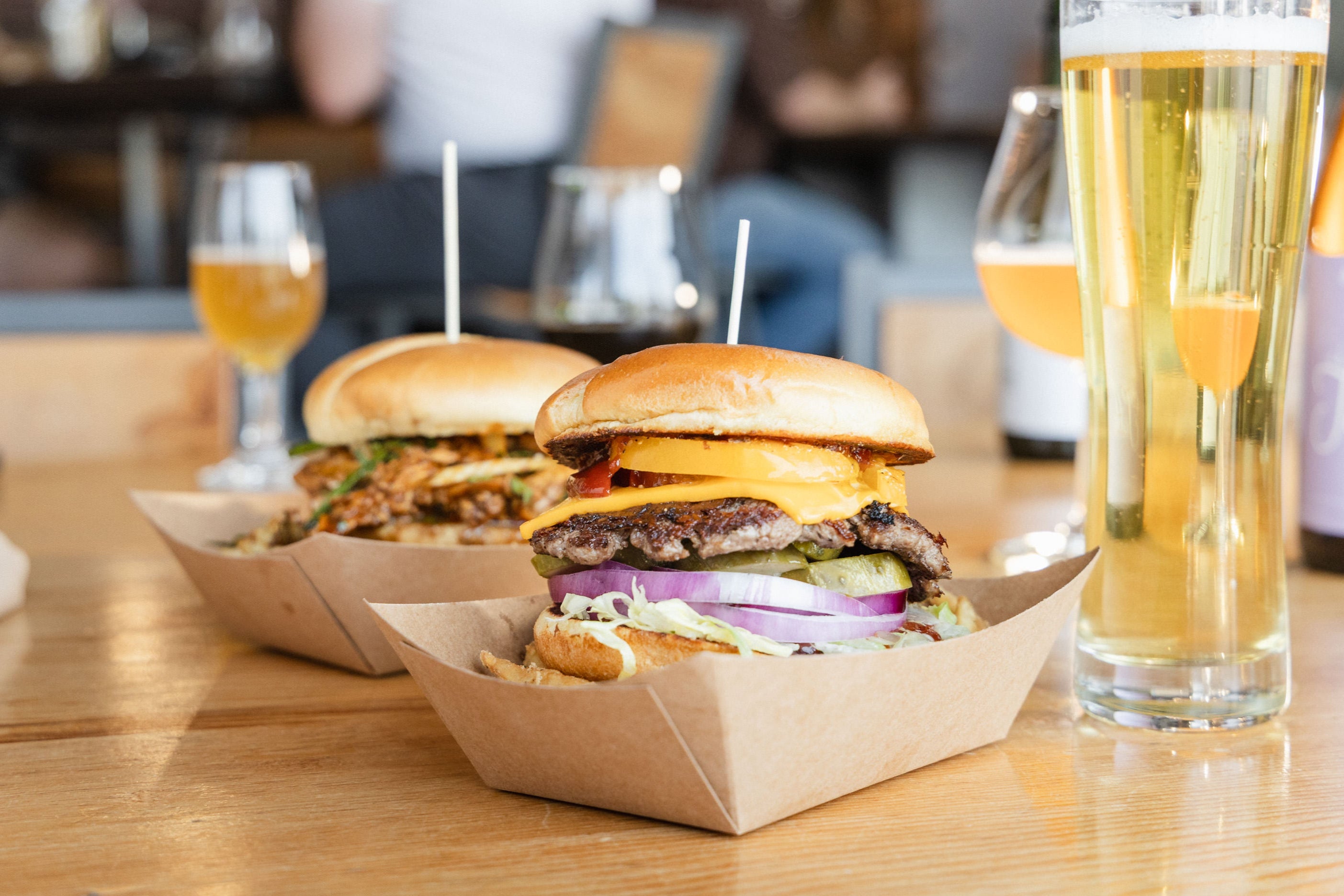 Two burgers in paper containers on a wooden table with glasses of beer.